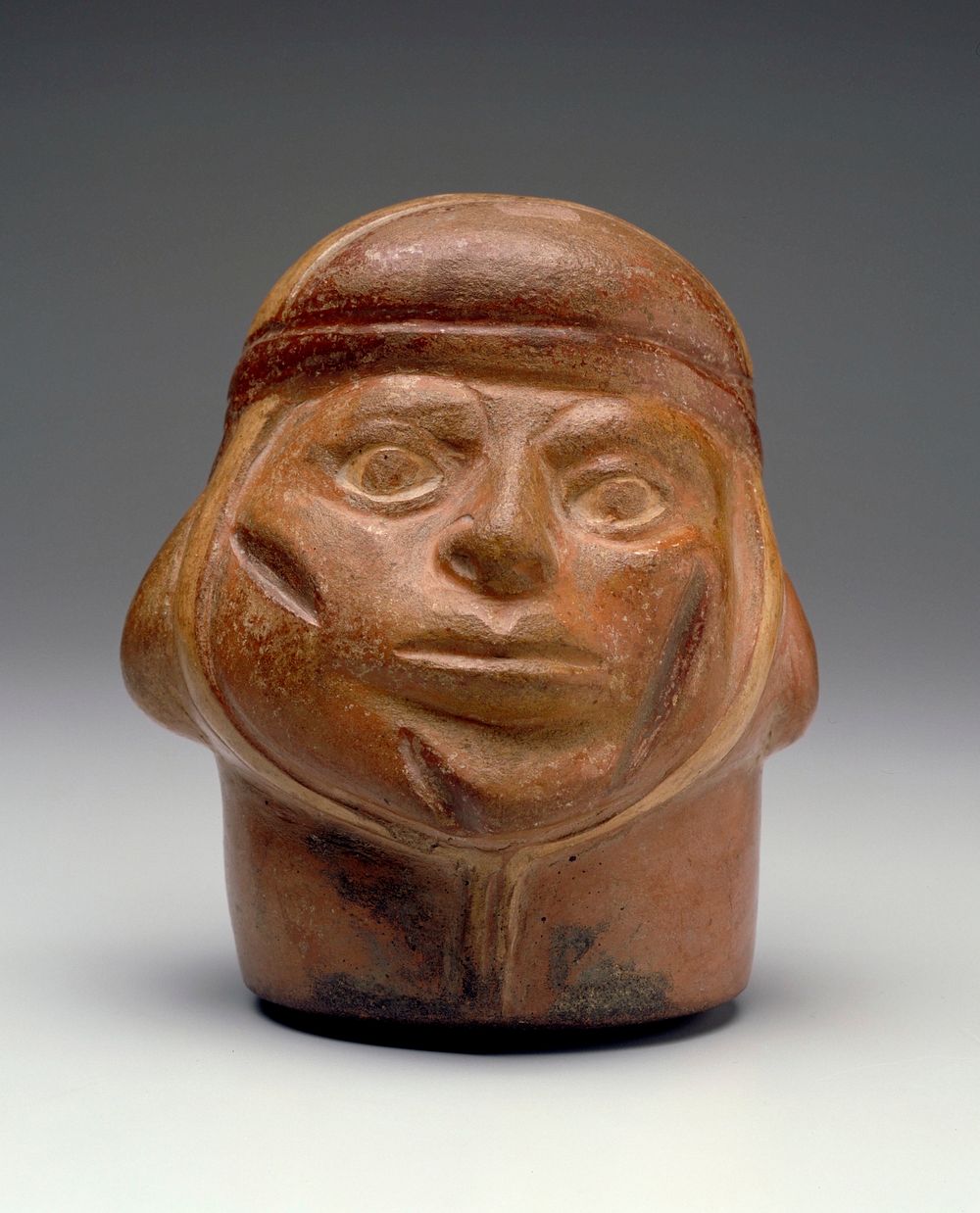 Portrait vase representing the head of warrior with three scars on his face. The head is closely wrapped in a scarf that…