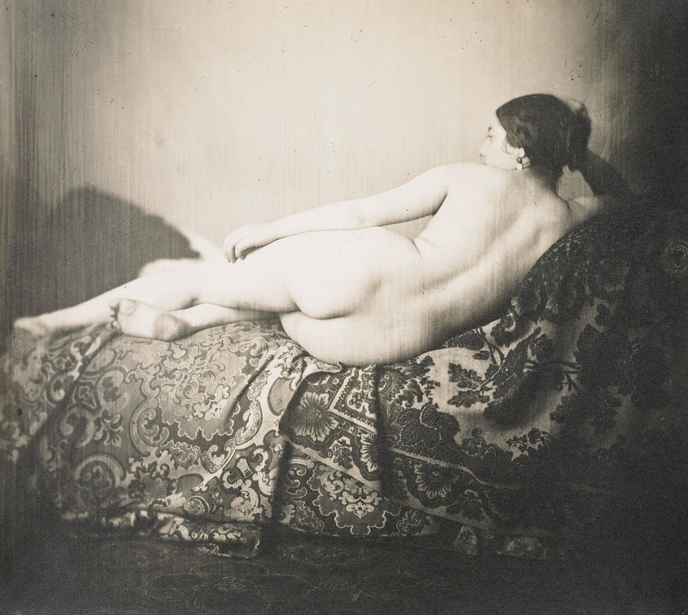 female nude lying on side on a couch facing away from the camera, head propped on hand; elaborately woven(?) fabric on…