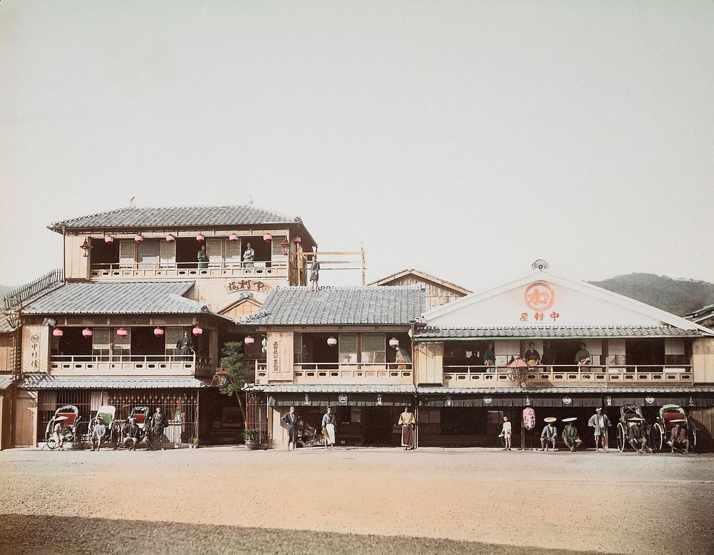 view across a wide street to a building with people standing on balconies, seated in rickshaws, standing on street in front…