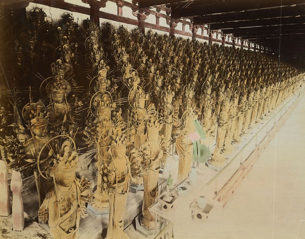 rows of many similar golden sculptures of Kannon in standing poses; green and pink flowers in vase near center foreground.…