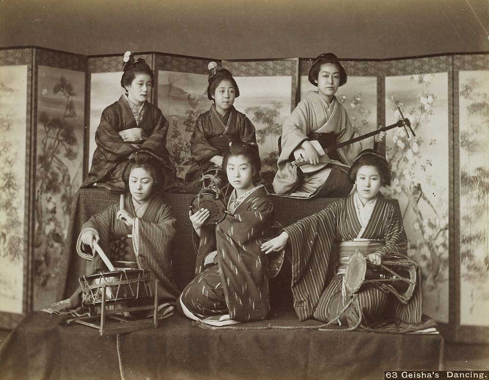 Six young women wearing traditional Japanese costumes, seated on two cloth-covered risers in front of folding screens; girl…