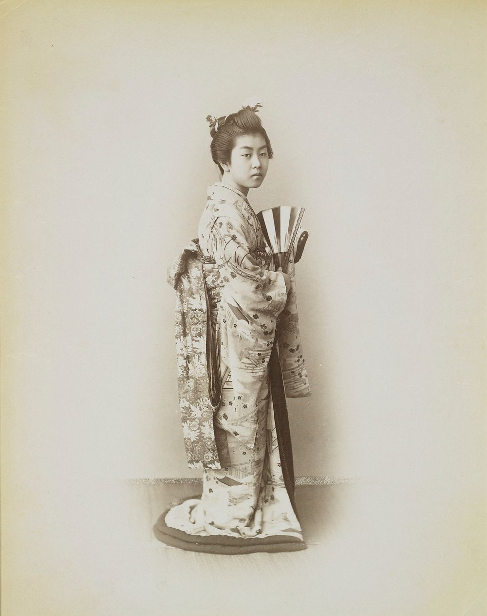 portrait of a standing girl wearing a patterned kimono with flowers and buildings and an obi with flowers, holding a striped…