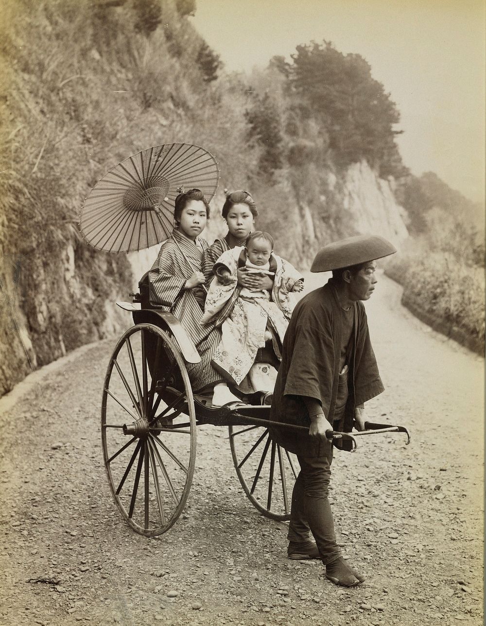two women wearing traditional Japanese costumes, seated in a rickshaw; woman at left holds a parasol; woman at right holds a…