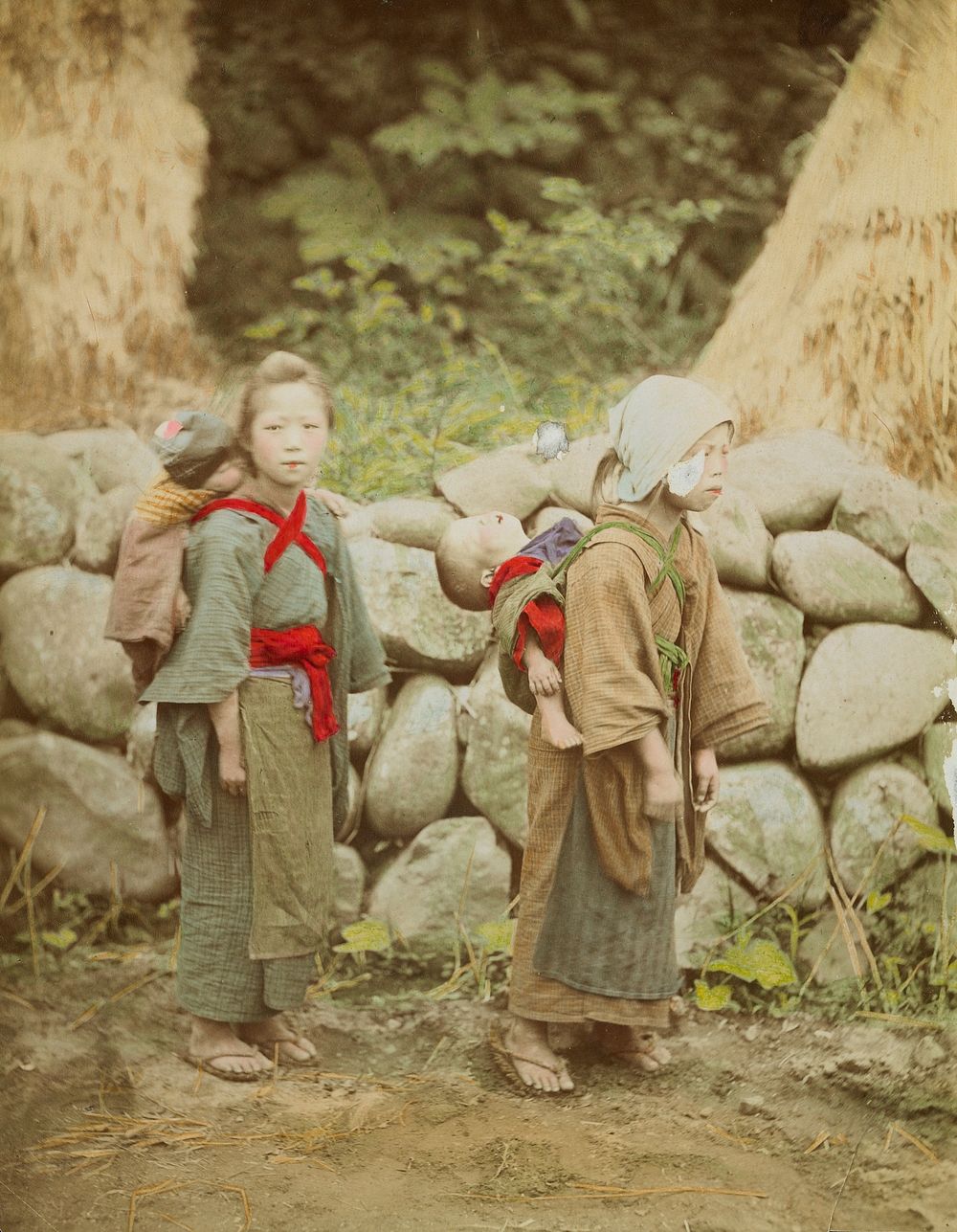 two girls with babies strapped to their backs, standing in front of a wall made of large rocks. Original from the…