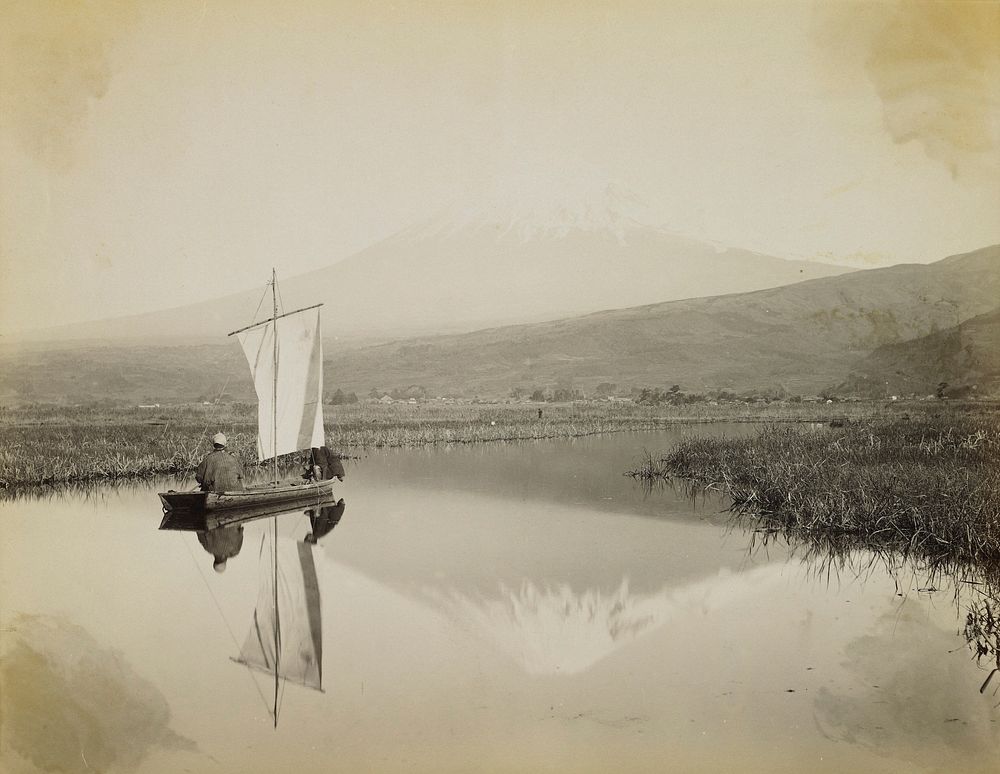 two figures in a small boat with a square sail on a calm body of water; snow-covered mountain in distance, reflected in the…