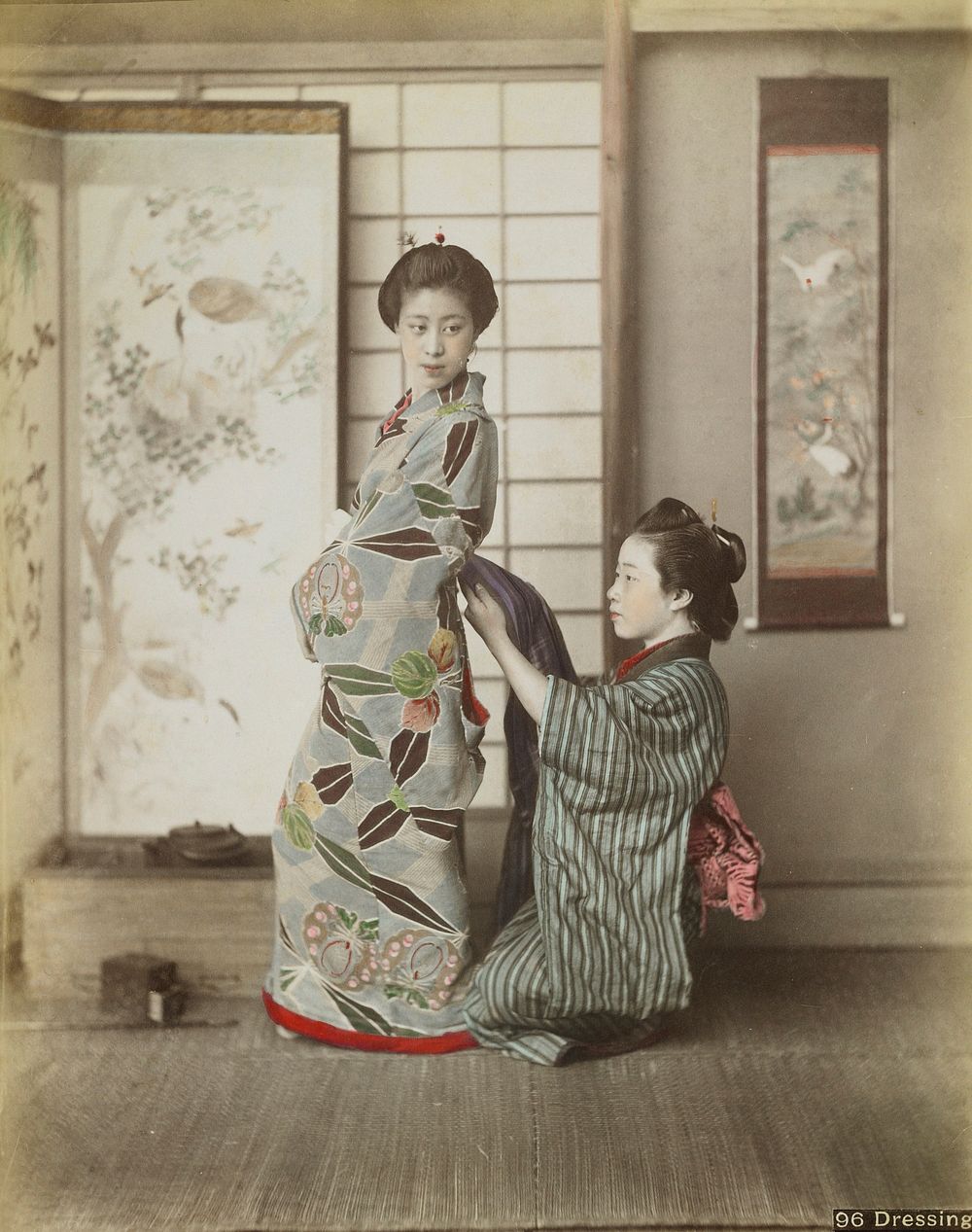 Two young women wearing traditional Japanese garments; kneeling woman at right wearing blue and grey striped kimono assists…