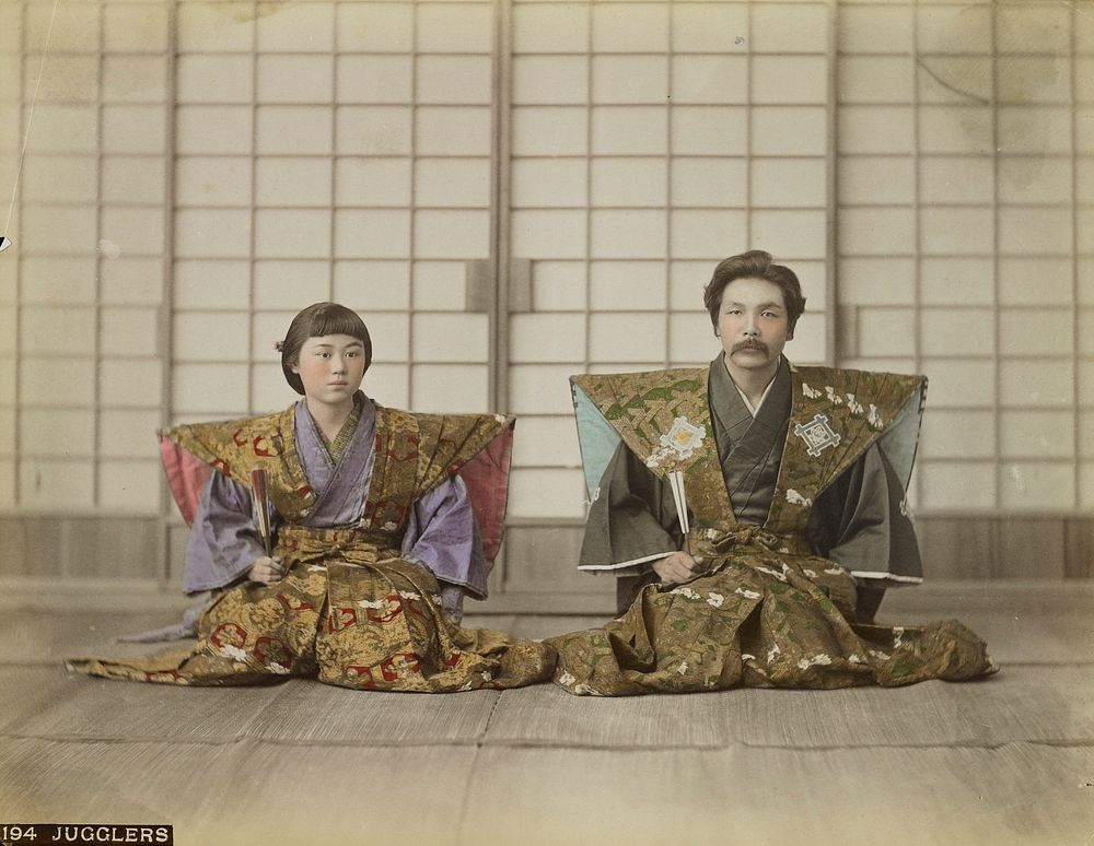 Seated young girl at left and man with moustache at right; both figures hold a closed fan in their PR hand and wear brocade…