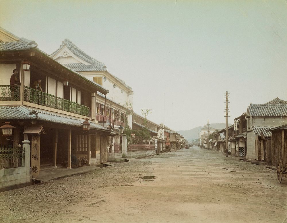 village scene in Japan; wide dirt road with buildings with tile roofs on each side--some with signs and banners in Japanese;…