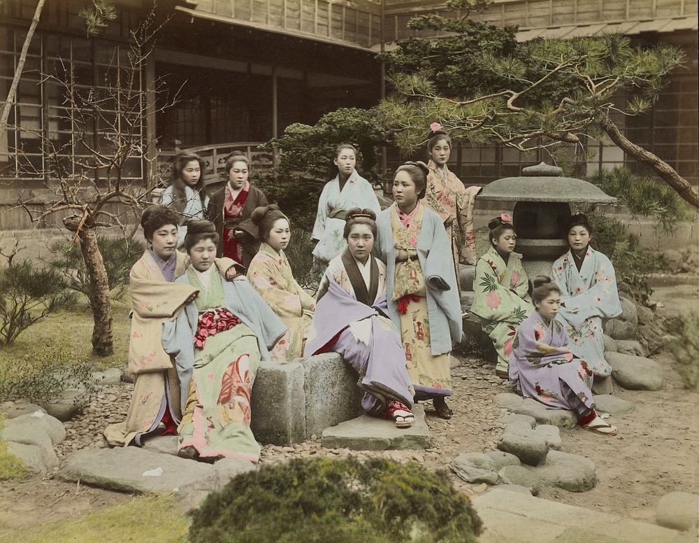 Twelve young women and girls in a Japanese garden; girls wear patterned, colorful kimonos; two girls have pink flowers in…