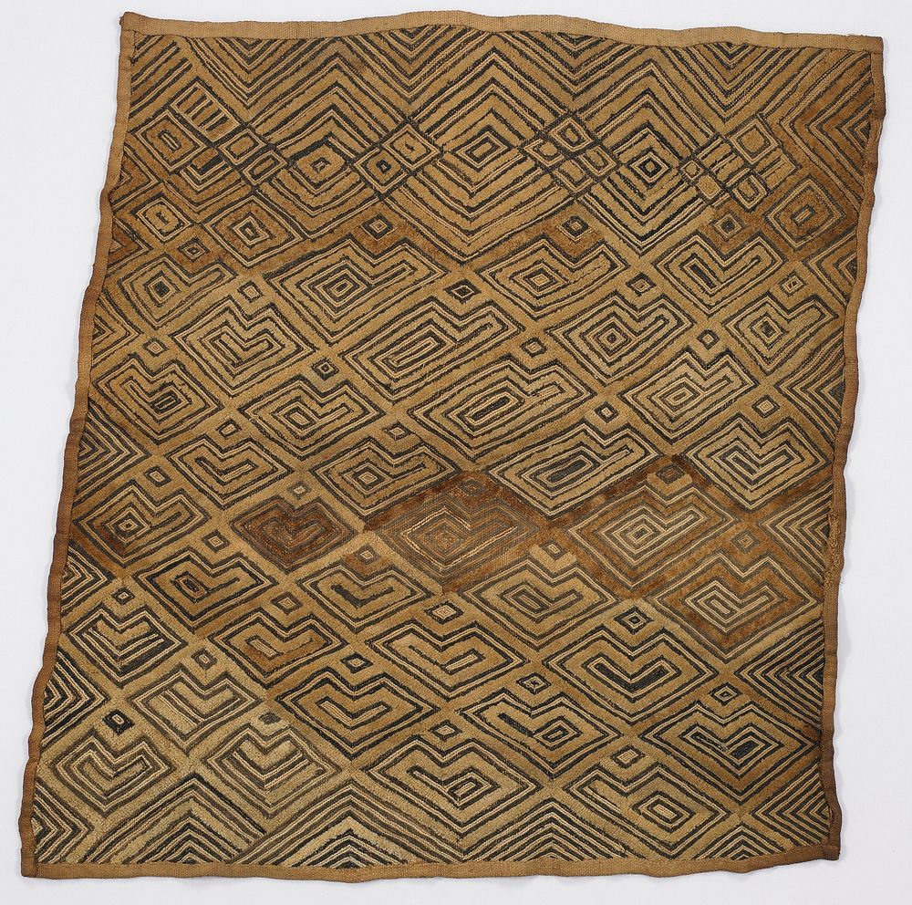 Tan clothwith P and L-shaped designs in dark fiber; zigzags and diamonds at one end; area of darker pile near center.…