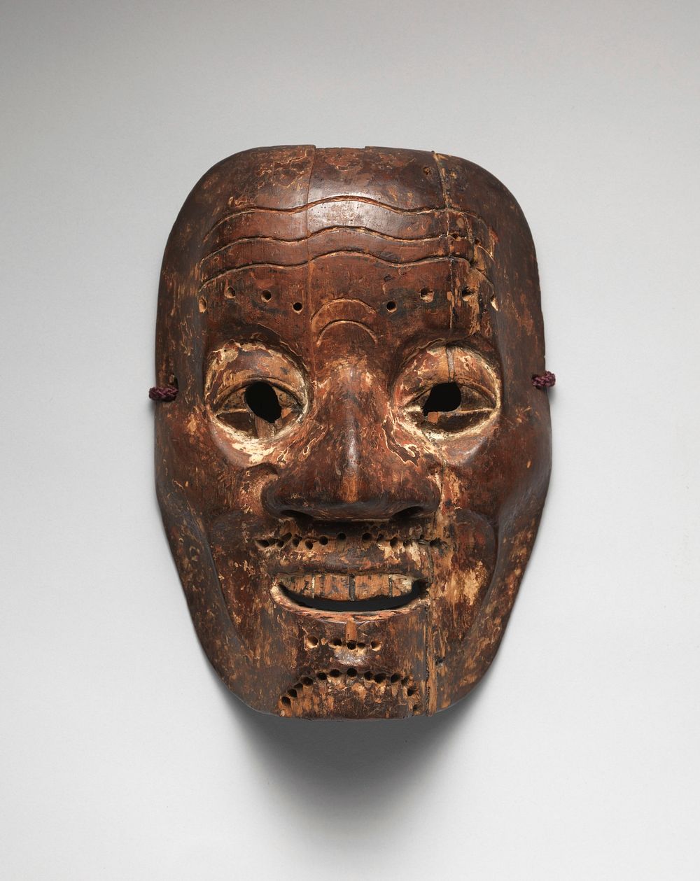 Mask of male face with large nose, round eyes with carved out eyeholes; narrow grin with top teeth visible; wrinkles in…