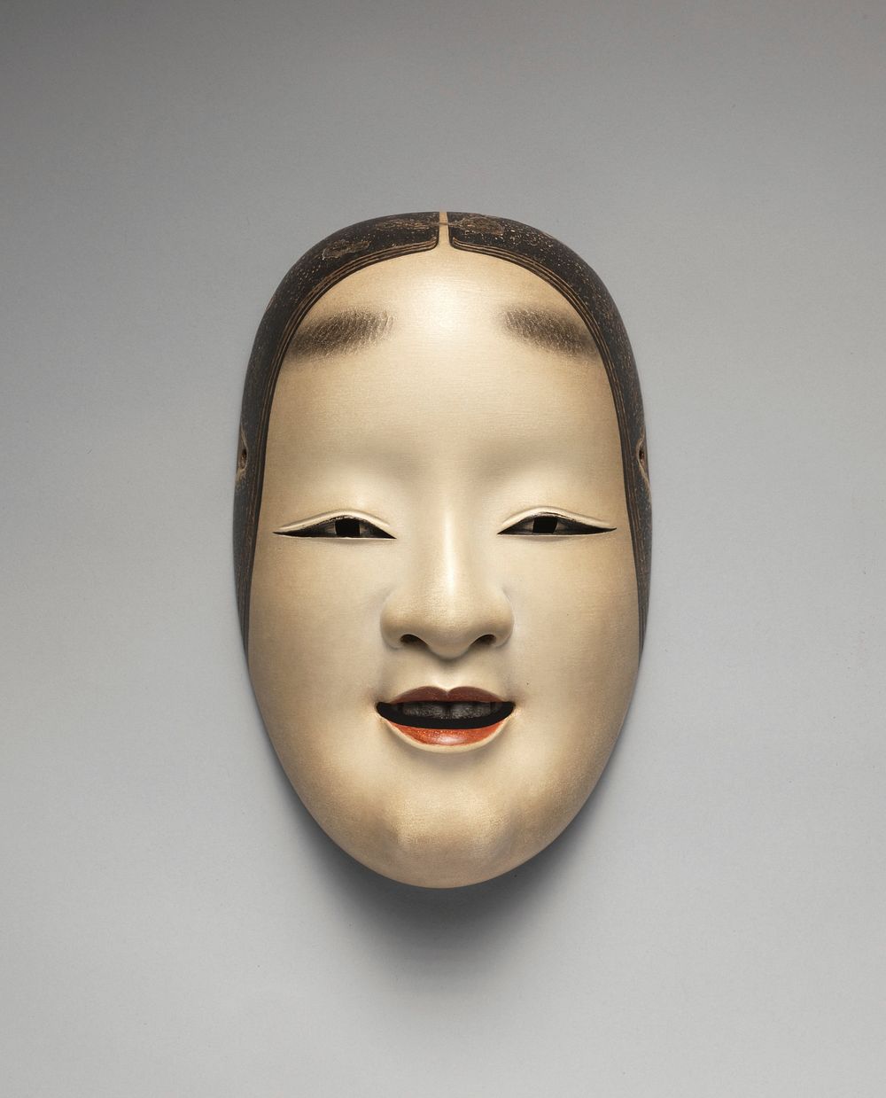 Mask of a slightly smiling female face; lips slightly parted with black teeth; eyes looking straight forward; painted…