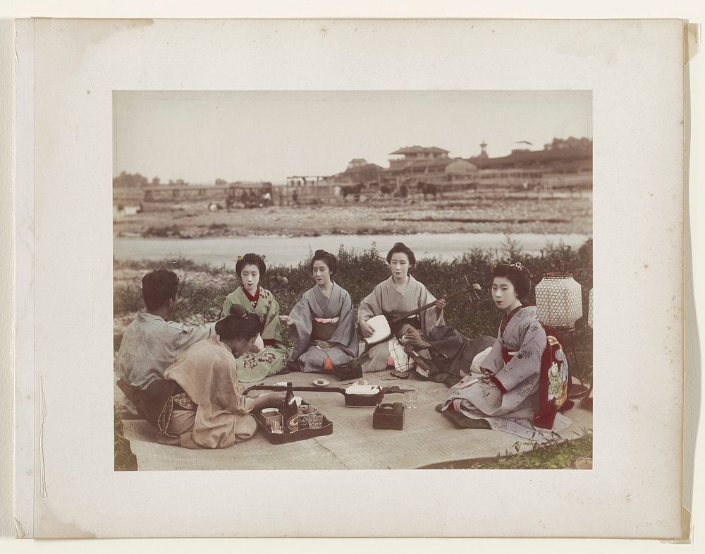 Small group of women with two men seated in semi-circle on woven reed blanket having a picnic; tray with food and beer, two…