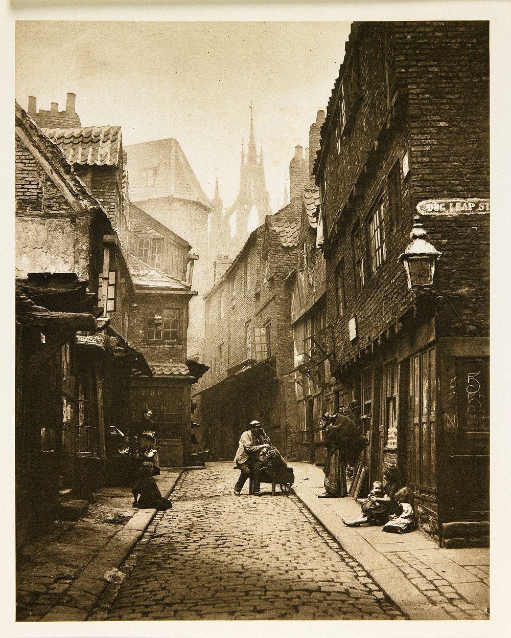 narrow, dark, brick street with tall building on both sides; shadowy castle towers visible in background; women and children…