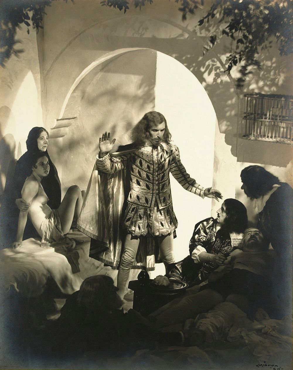 staged shot of man in period costume beneath arch surrounded by two women, one nude, on proper left, three men on proper…