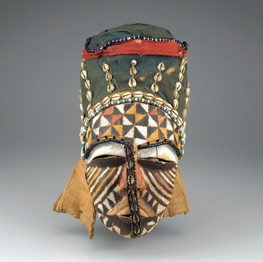 small face with wide slit eyes; face decorated with white, tan and brown lines and triangles; cloth band with shells down…