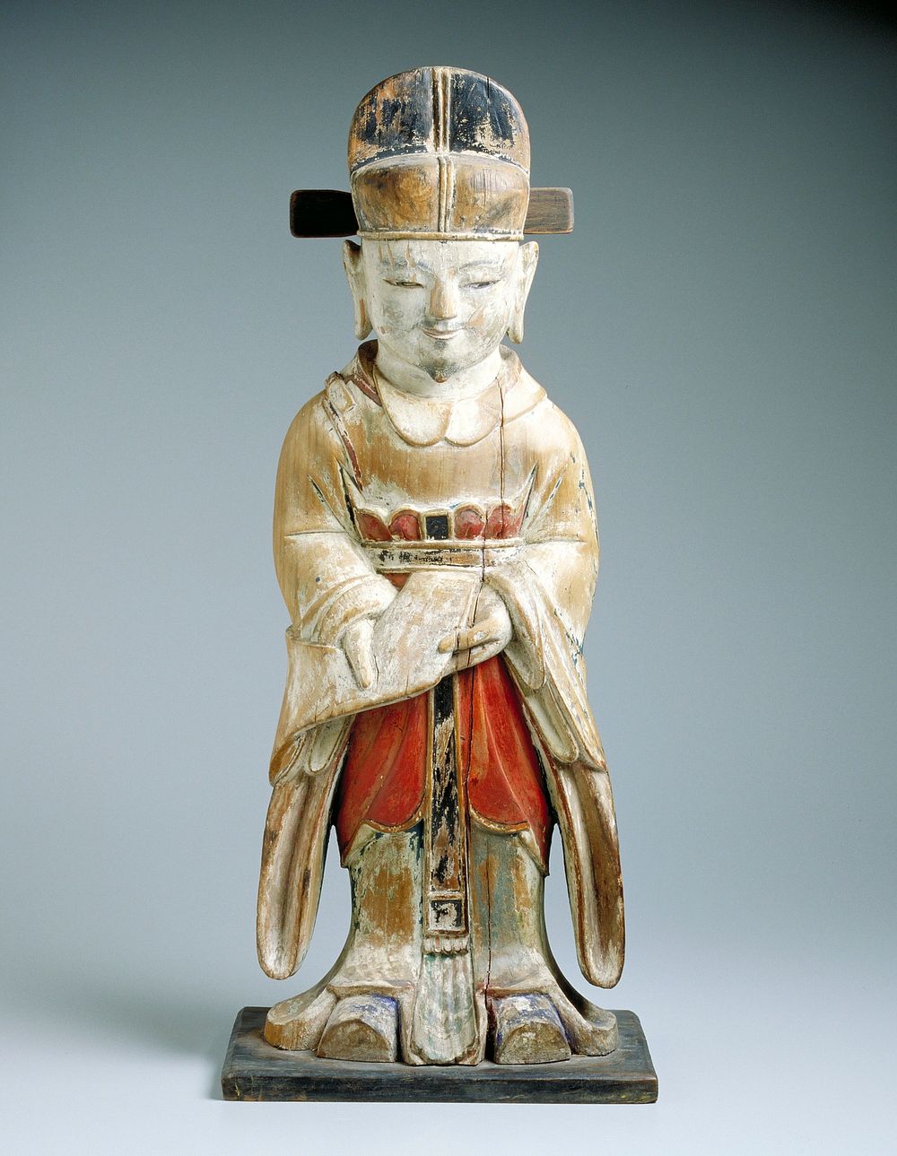 polychrome wood statue of a Confucian scholar standing, holding a book (?) downward at waist toward the viewer, wearing a…