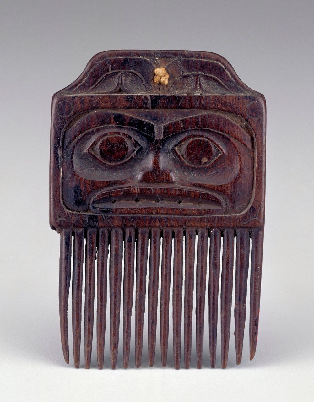 comb topped with face with slightly sunken eye sockets and wide mouth on one side; lightly incised, abstracted face with…
