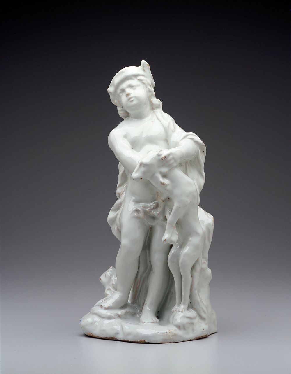 faience figure of a nude youth wearing a rustic hat, holding a standing dog by its neck; covered by a viscous white glaze.…