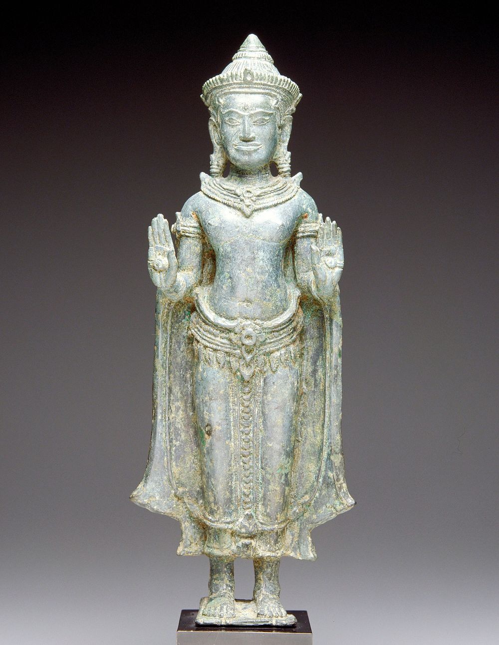 standing Buddha wearing cape; both hands raised in abaya mudra; wearing caplike crown, earrings and necklace; attached to…