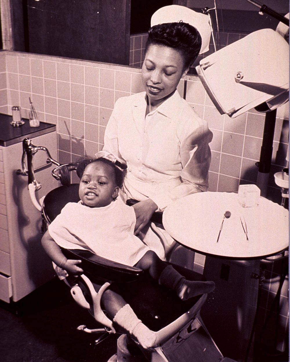 Child sitting in dental chair. An African American little girl is sitting in a dental chair with a dental assistant standing…