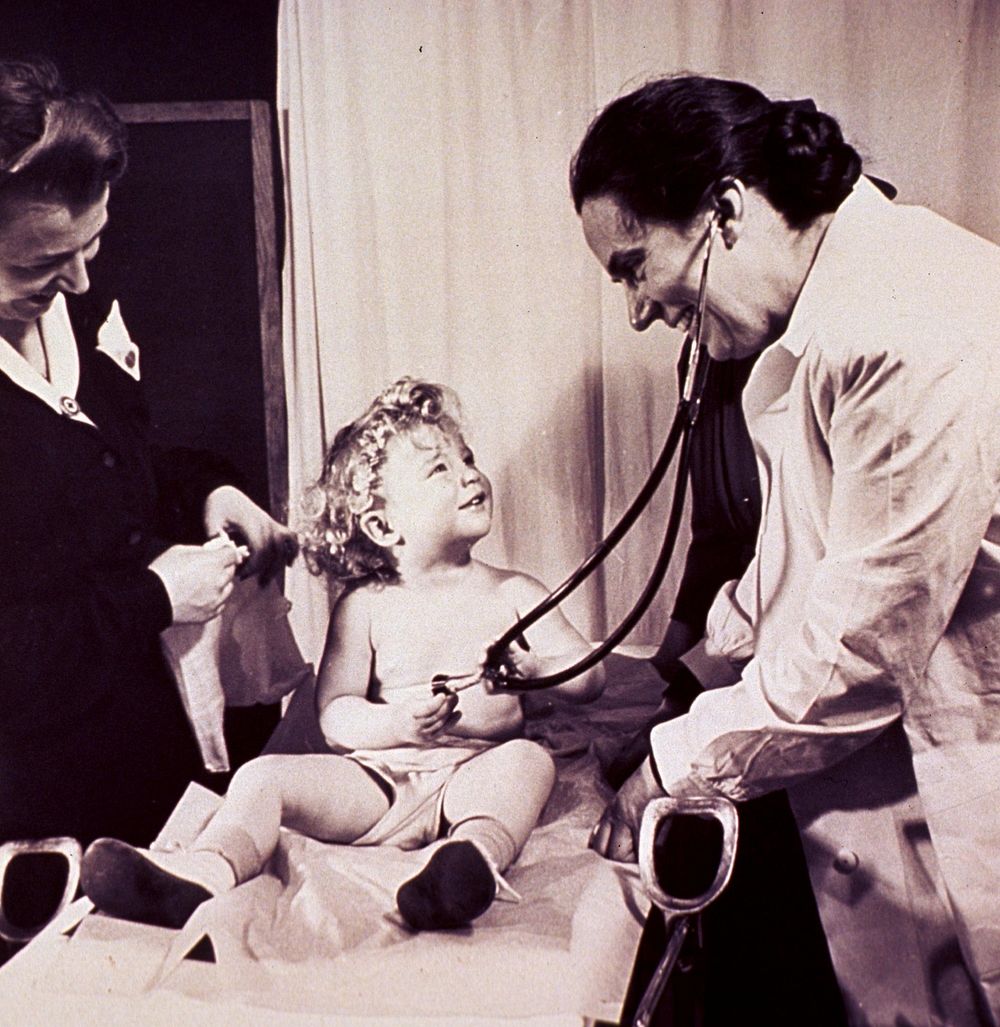 Public health nursing Medical exam. A child, sitting on an obstetrical table, is playing with a physician's stethoscope.…