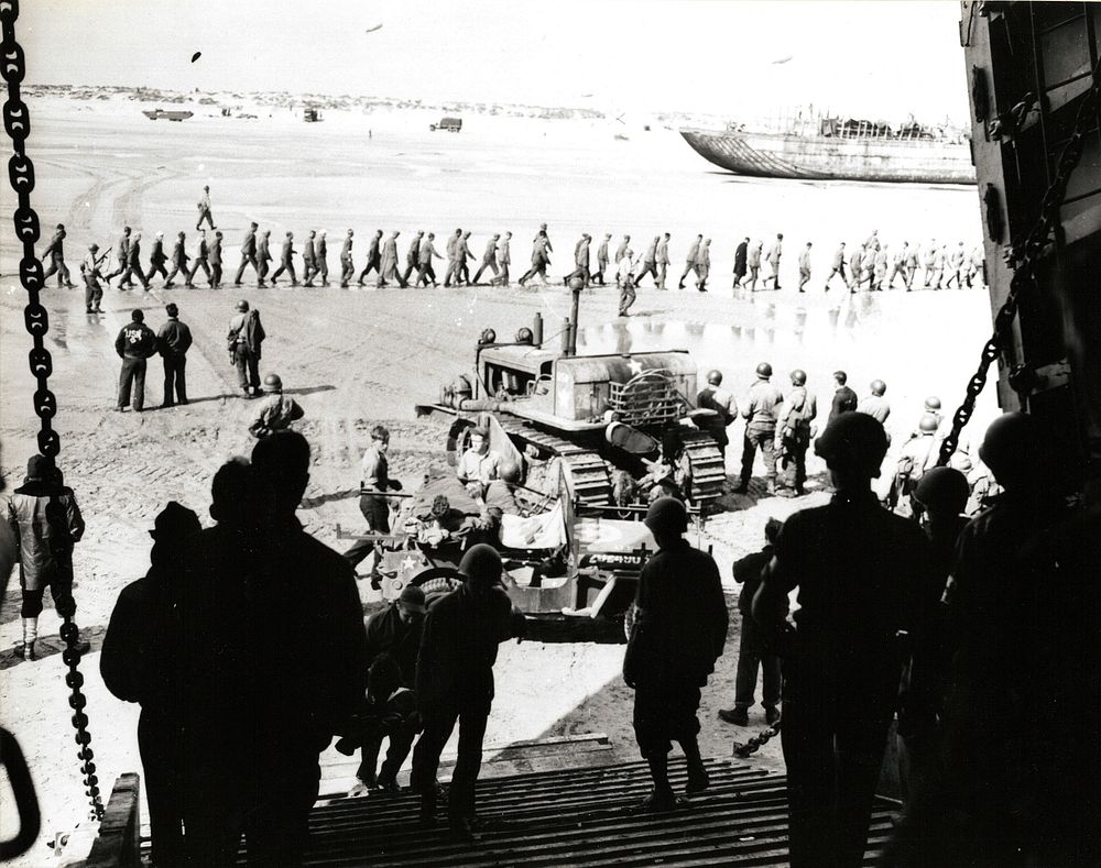 Casualties are brought aboard an LST for removal from the beachhead. In the background are German prisoners under guard.…