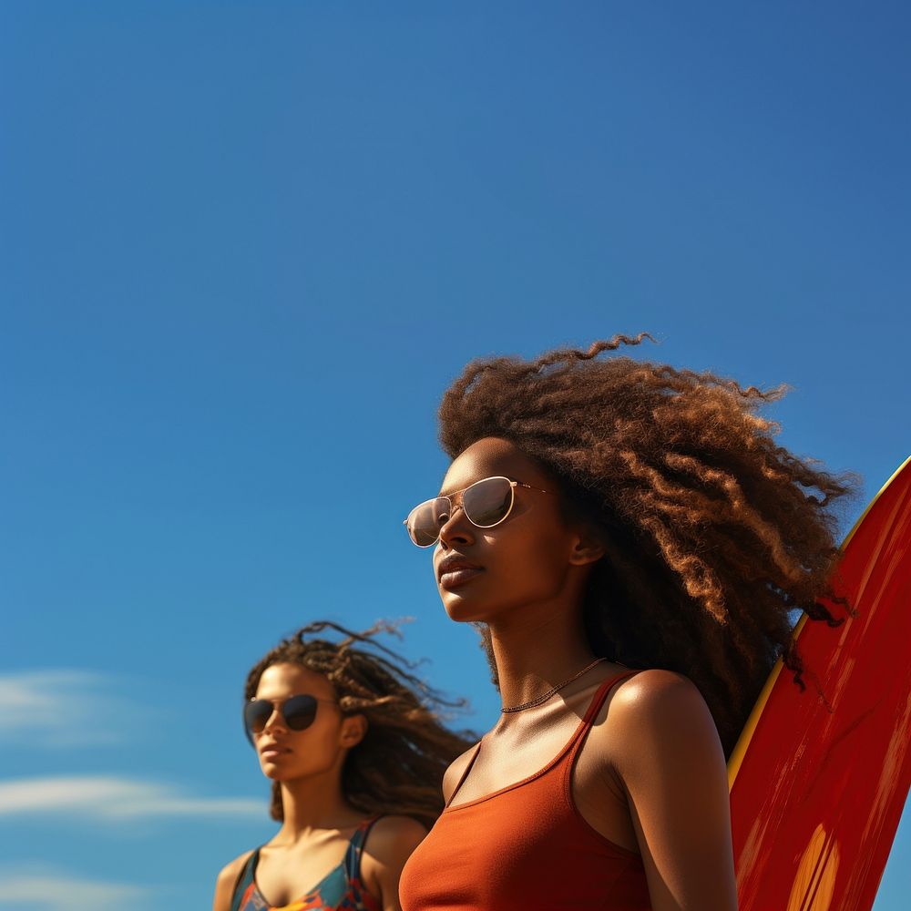 Photo of a black woman carrying surfboard with her friend on the beach, on a blue sky, sunny day.  