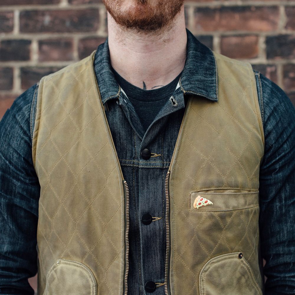 A bearded man who is standing in front of a brick wall is wearing a pizza pin on his distressed vest.