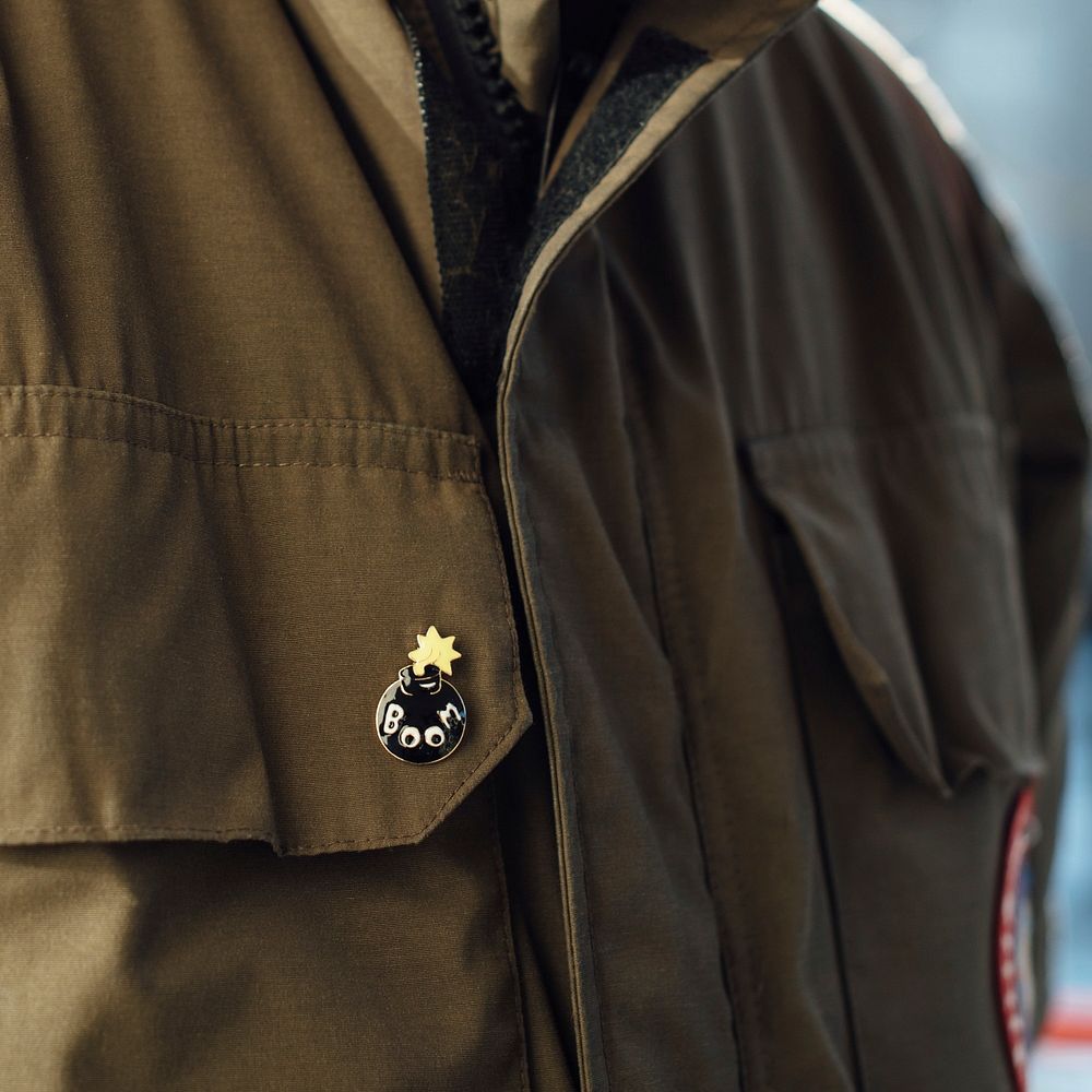 A man is wearing a trendy enamel bomb pin on the pocket of his military green shirt.