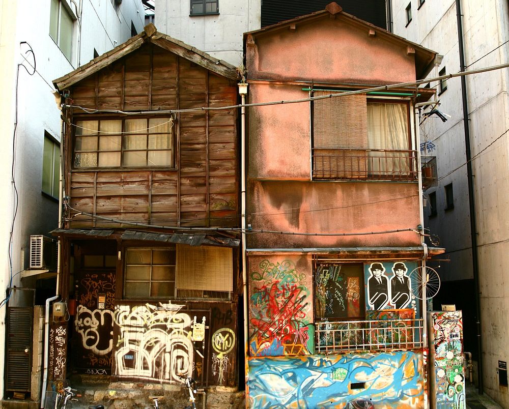 Two tall ramshackle wooden buildings covered in spray paint and street art stand surrounded by taller concrete buildings.