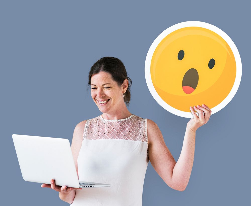 Woman holding a surprised emoticon and using a laptop