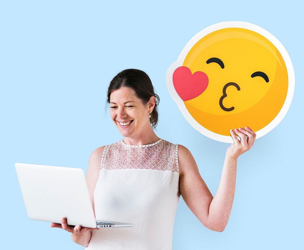 Woman holding a kissing emoticon and using a laptop