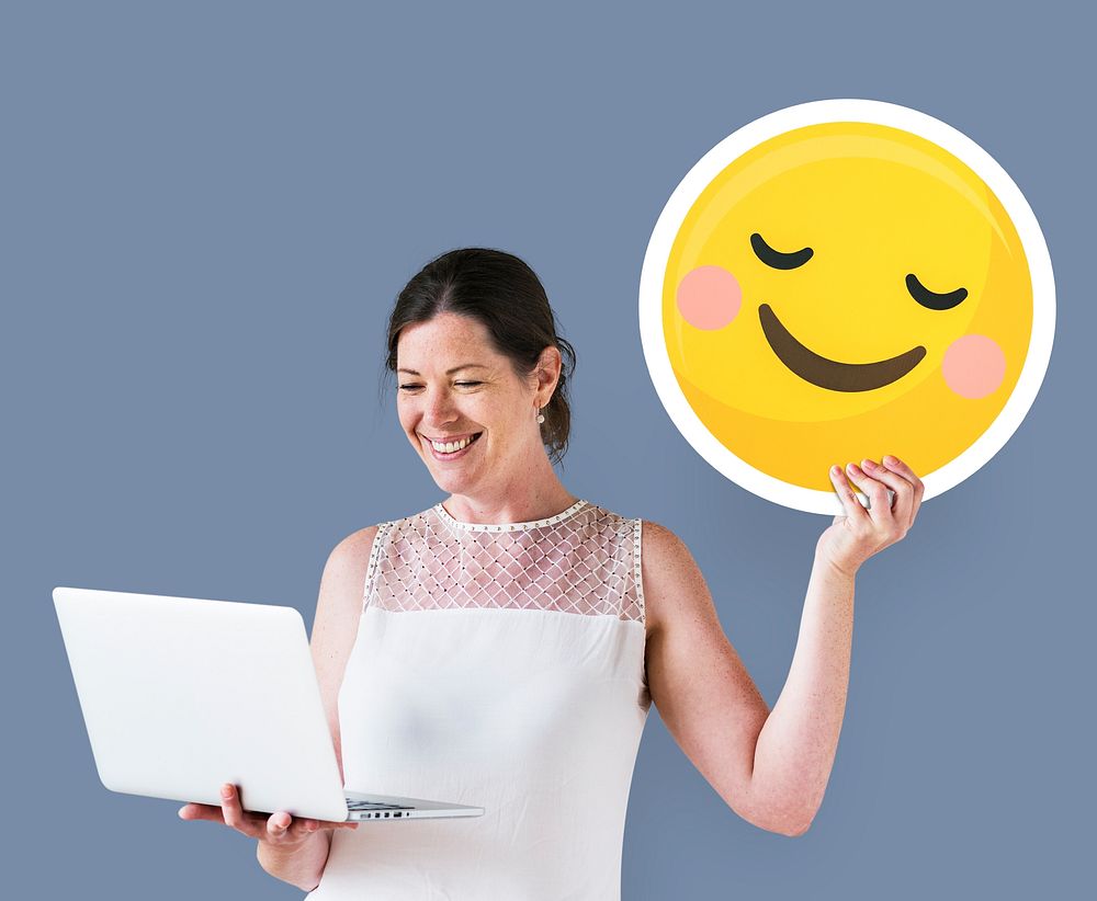 Woman holding a blushing emoticon and using a laptop