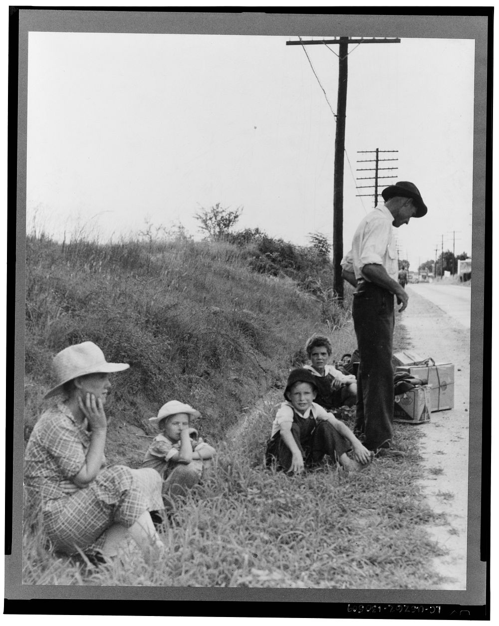A hitchhiking family waiting along the highway in Macon, Georgia. The father repairs sewing machines, lawn mowers, etc. He…