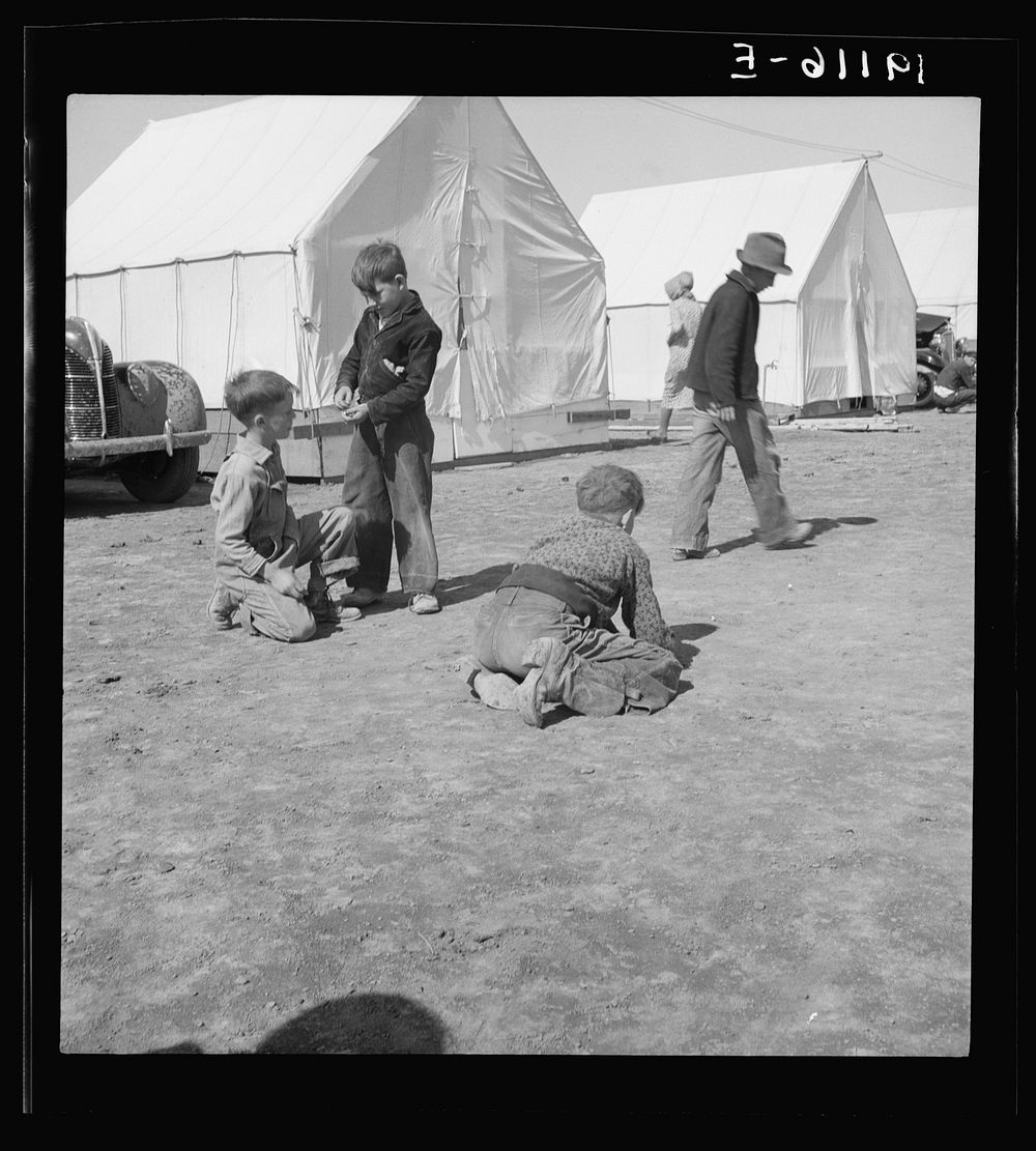 Migrant children in Farm Security Administration (FSA) migratory labor camp. Note type of boots worn by boy shooting marbles…