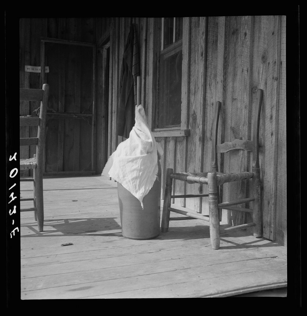 Pottery butter churn on porch of  tenant family. The churn is covered with a cloth to keep the flies out. Note chair seat of…