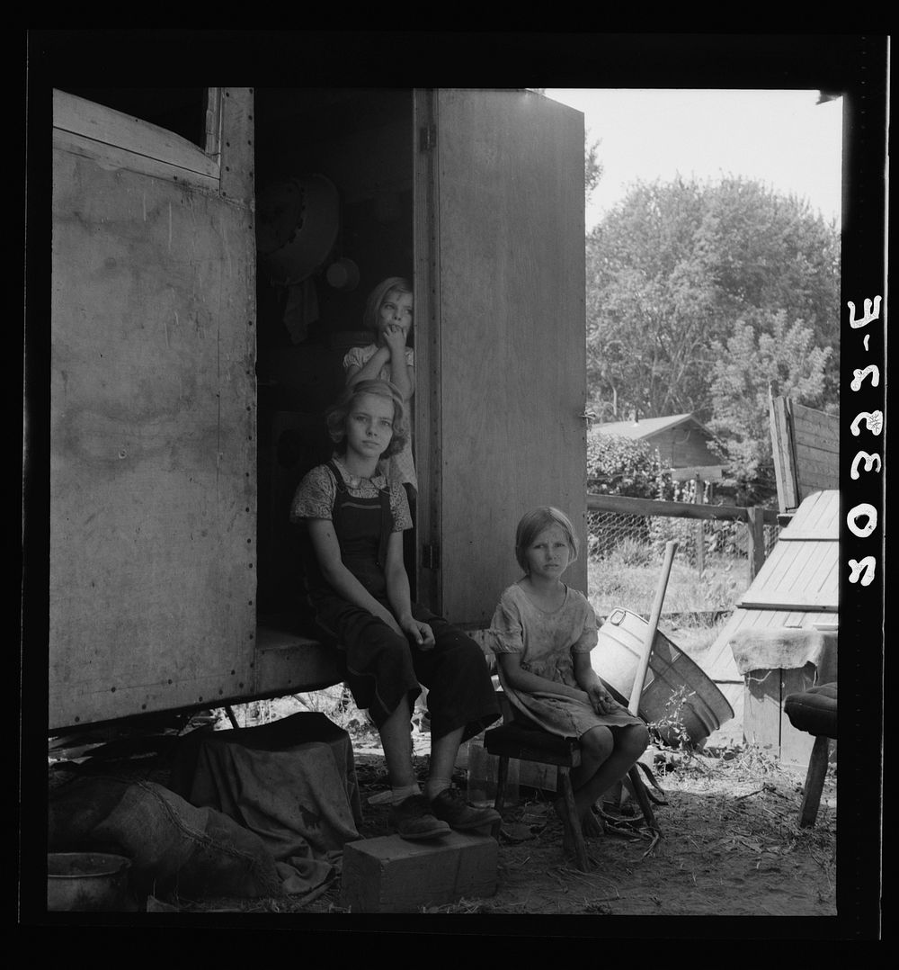 The oldest girl seated in the doorway of the house trailer cares for the family. Yakima Valley, Washington. Sourced from the…