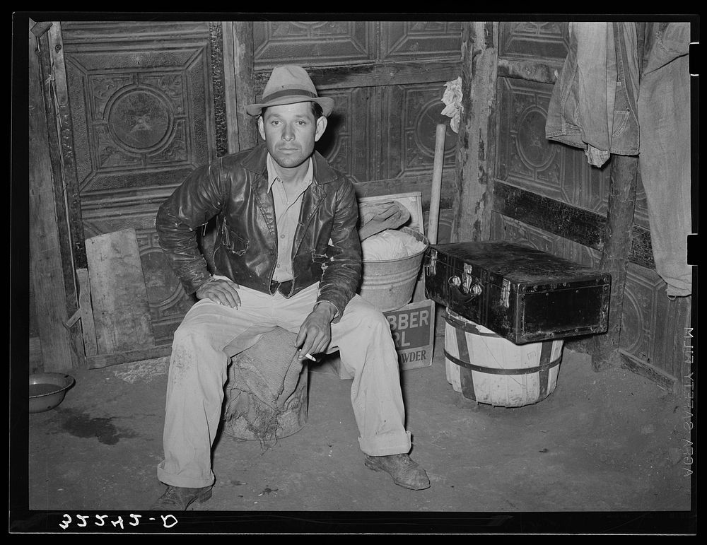 Mexican man in corner of one-room dwelling. Notice dirt floor and siding of house which is constructed of discarded tin…