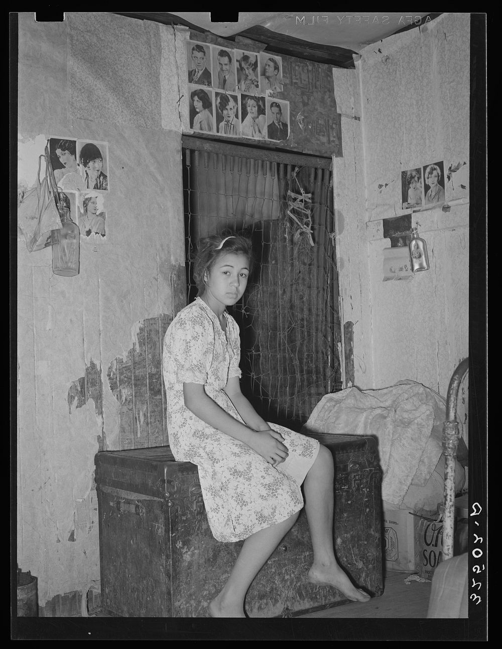 Mexican girl sitting in front of bedspring which formed partition between the family's living quarters and those of a man…