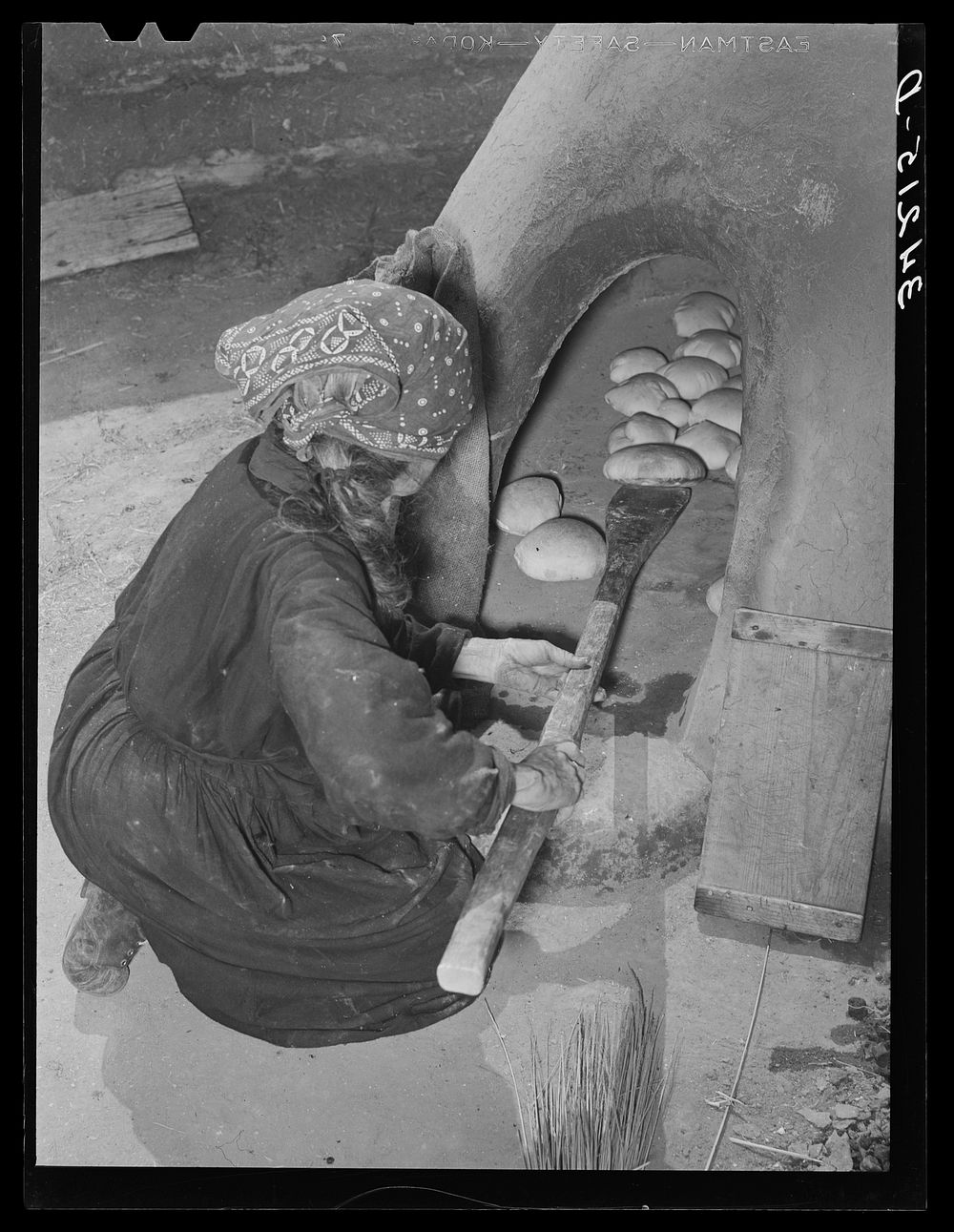 Spanish-American woman removing baked bread from outdoor earthen oven by means of a long wooden paddle. Near Taos, New…