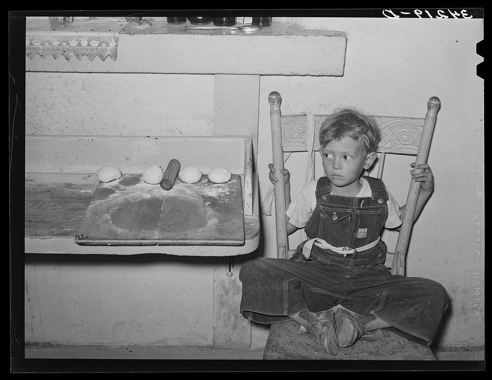 Spanish-American boy sitting in chair by tortilla board. Near Taos, New Mexico, Taos County by Russell Lee