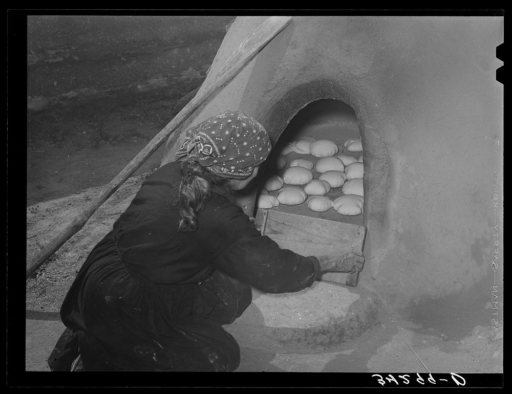 Spanish-American woman putting boards over entrance of outdoor oven to retain heat for baking bread. Near Taos, New Mexico…