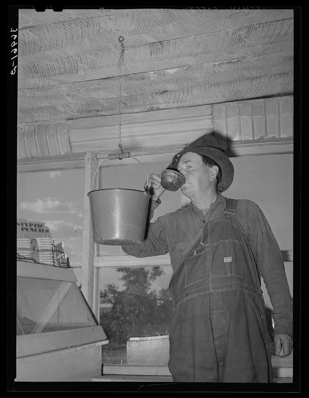 Farmer drinking water from bucket which hangs in the general store. Pie Town, New Mexico by Russell Lee