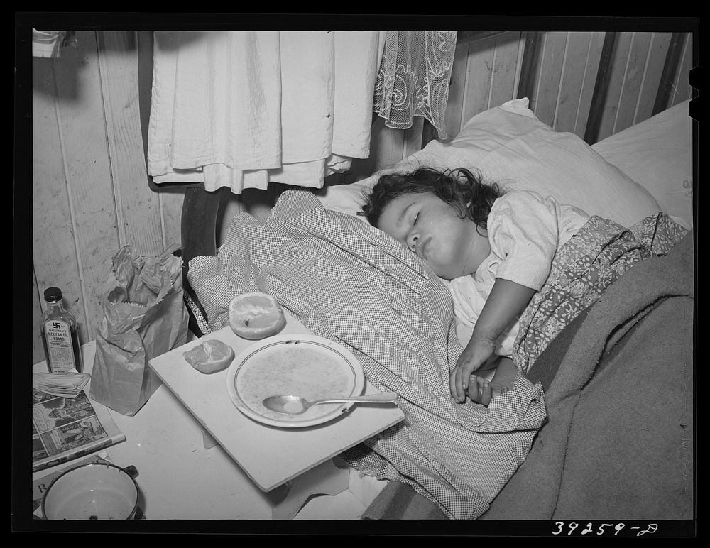 Sick child in bedroom of house in the Mexican section. Seven children and two adults sleep in two small rooms and in two…