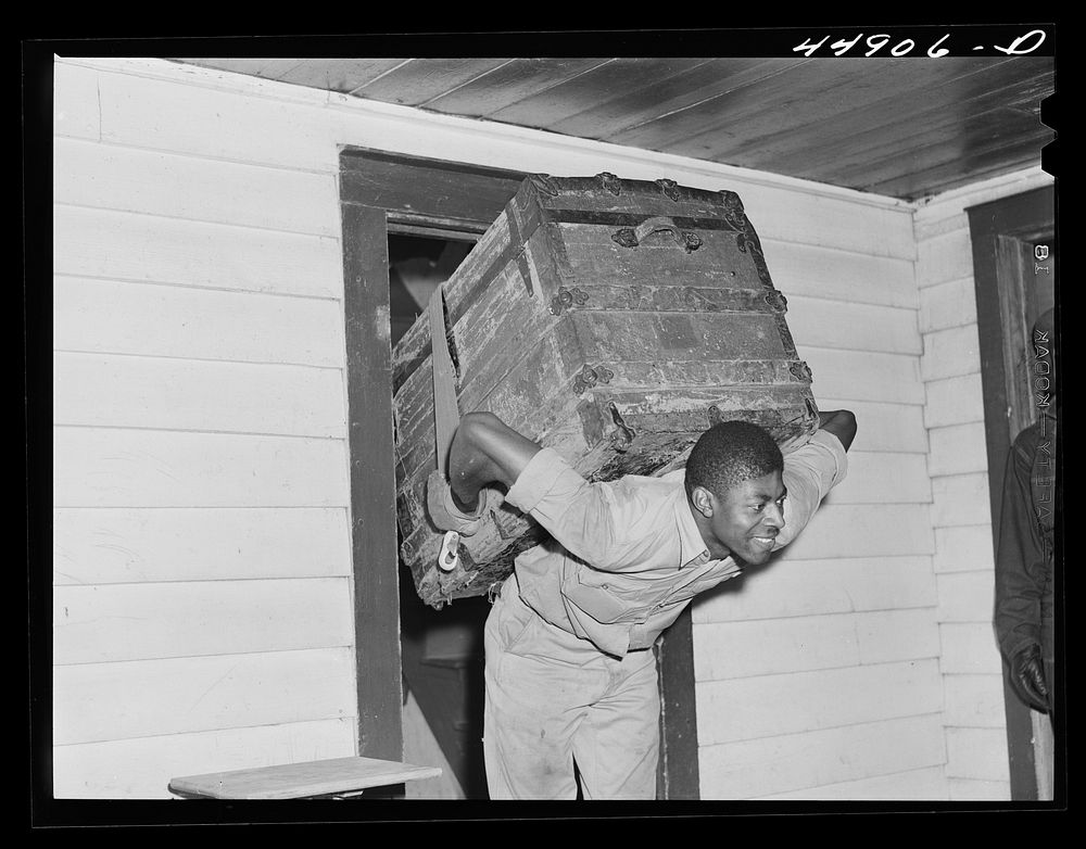 CCC (Civilian Conservation Corps) boy carrying a trunk out for a family who is moving out of the area taken over by the Army…