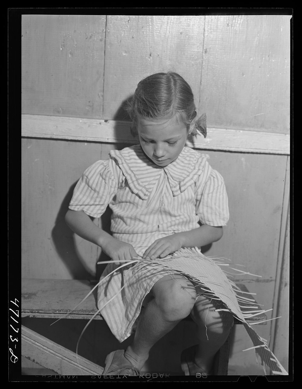 French village, a small settlement on Saint Thomas Island, Virgin Islands. Little girl weaving palm leaf mat to be sold at…