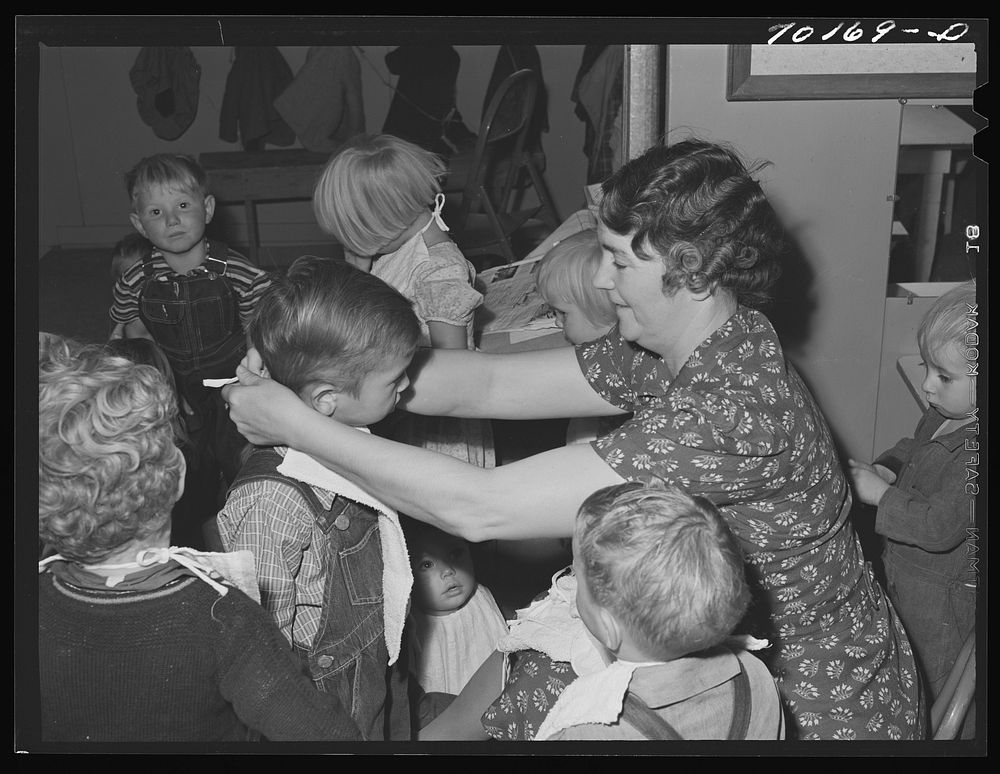 WPA (Work Projects Administration) teacher at the nursery school puts on bibs of her charges at the FSA (Farm Security…