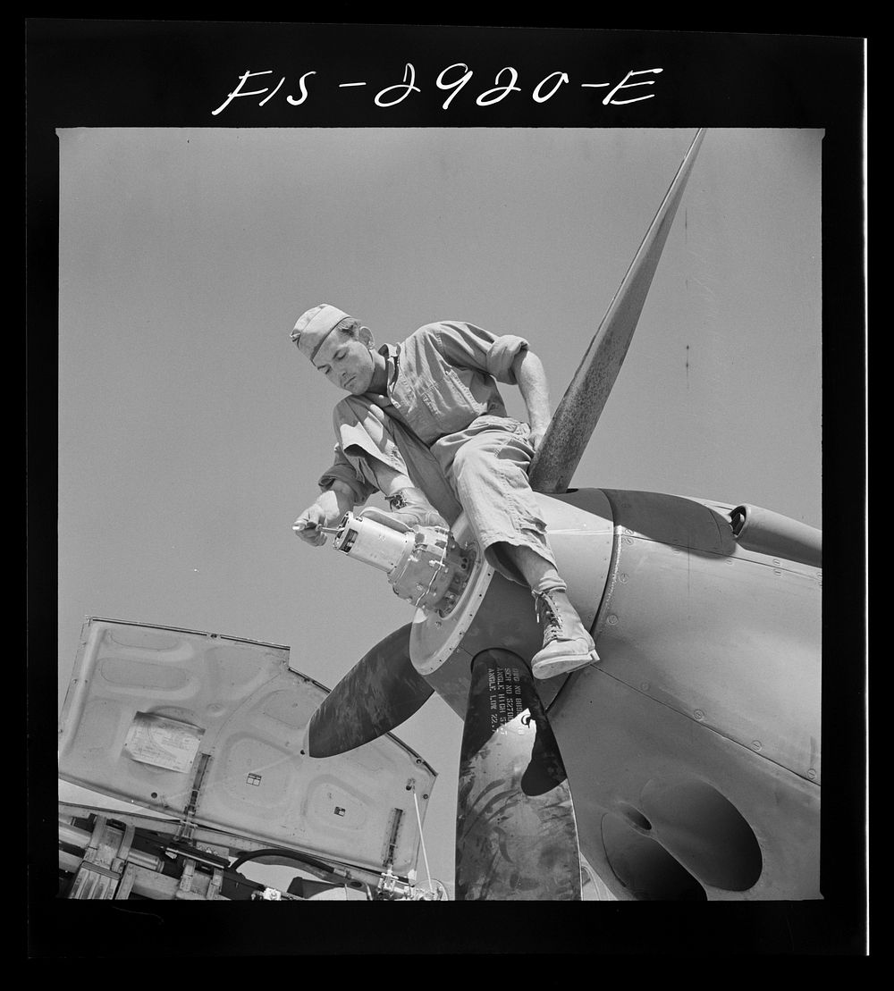Repair man perched upon propeller hub of airplane. Lake Muroc, California by Russell Lee