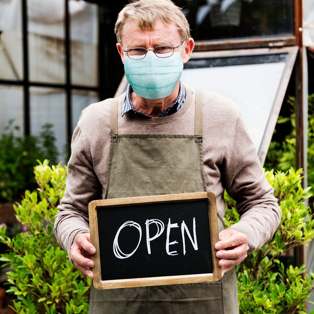 Senior shop owner in mask with open sign at plant nursery